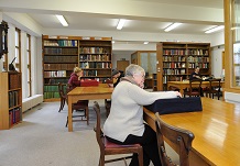 Researchers sitting at tables in a lights, book-lined study room, with documents on book rests in front of them